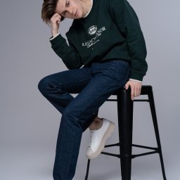 Aleksandra Linke - Młody mężczyzna w ciemnozielonej bluzie z logo, ciemnych jeansach i białych sneakersach, siedzący na czarnym stołku barowym na jednolitym szarym tle.