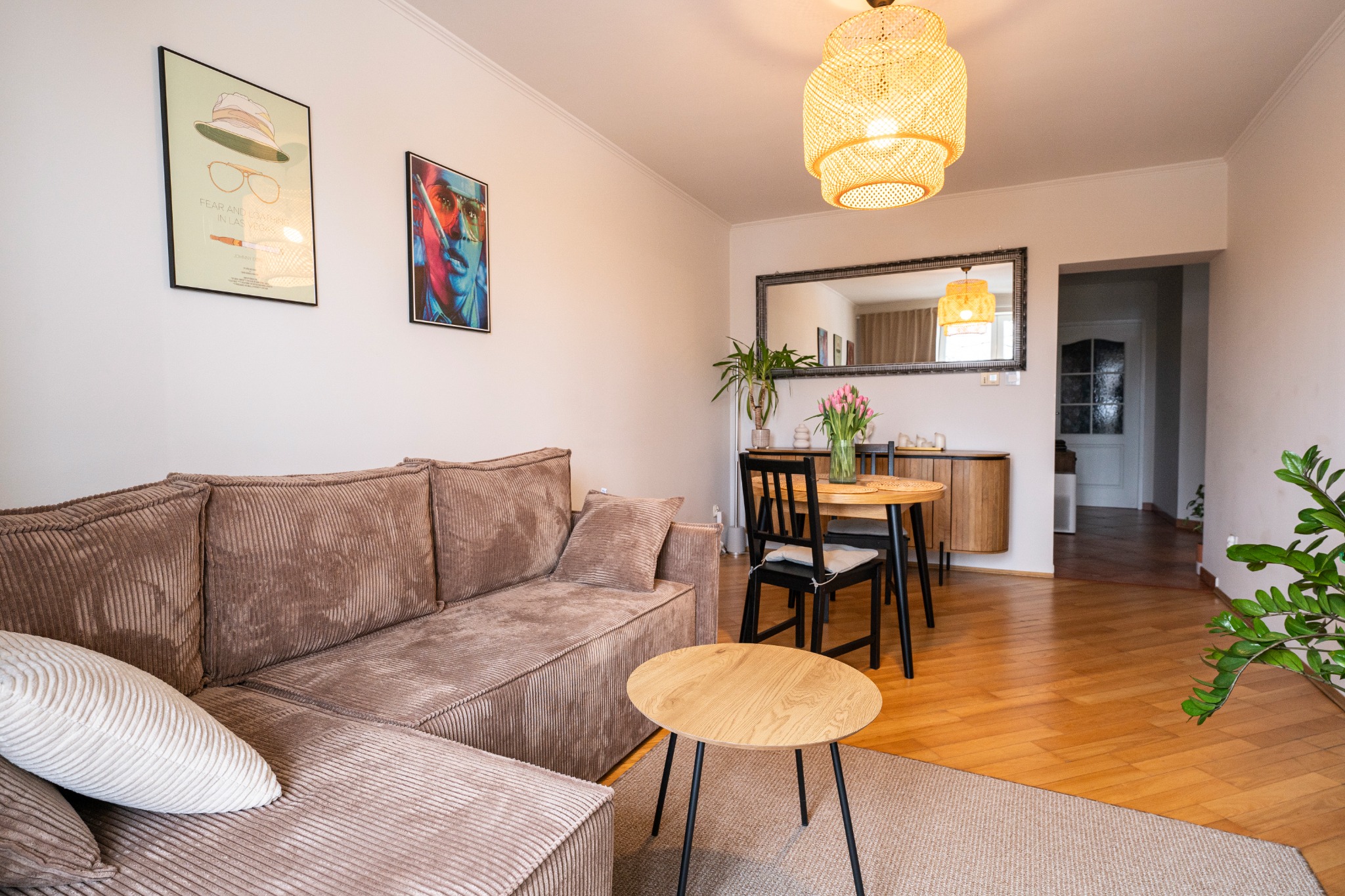 Jasne wnętrze salonu z brązową sofą narożną, okrągłym stolikiem kawowym, drewnianym stołem z czarnymi krzesłami, lustrem w czarnej ramie i dekoracyjnymi plakatami na ścianie.