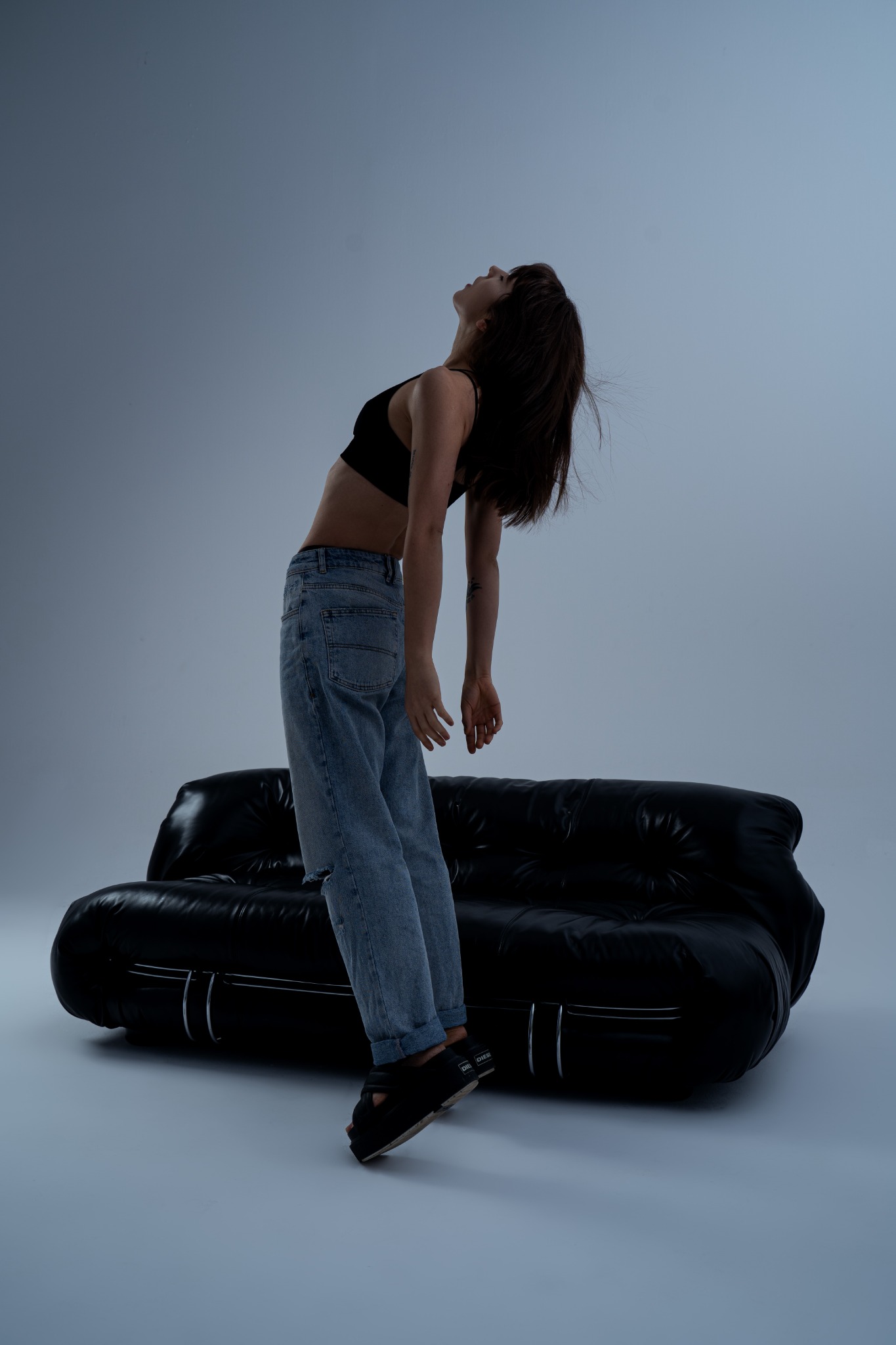 Kobieta w dżinsach i sportowym topie odchyla się do tyłu na czarnej skórzanej sofie, patrząc w górę na jasnym, neutralnym tle studyjnym.