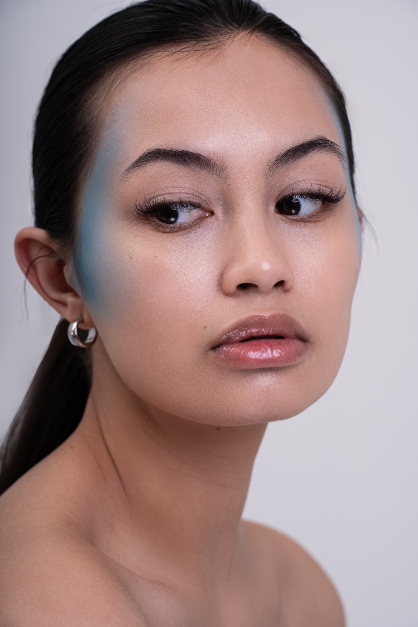 Portret kobiety z delikatnym makijażem w odcieniach nude i akcentem błękitu na skroniach, patrzącej w bok, z srebrnym kolczykiem kołem.