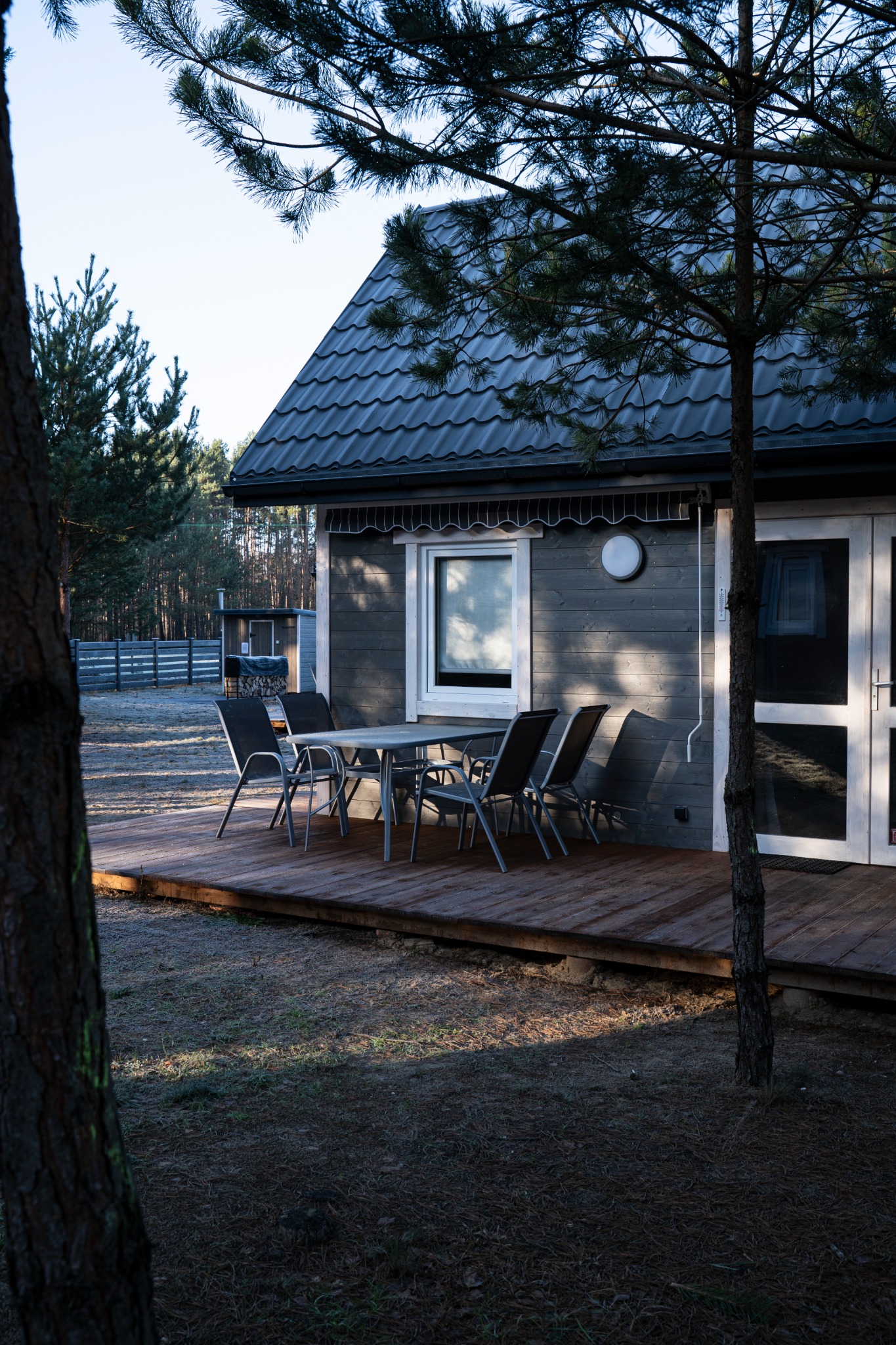 Szary drewniany dom z tarasem i stołem z krzesłami, widok przez gałęzie drzewa, w tle las.