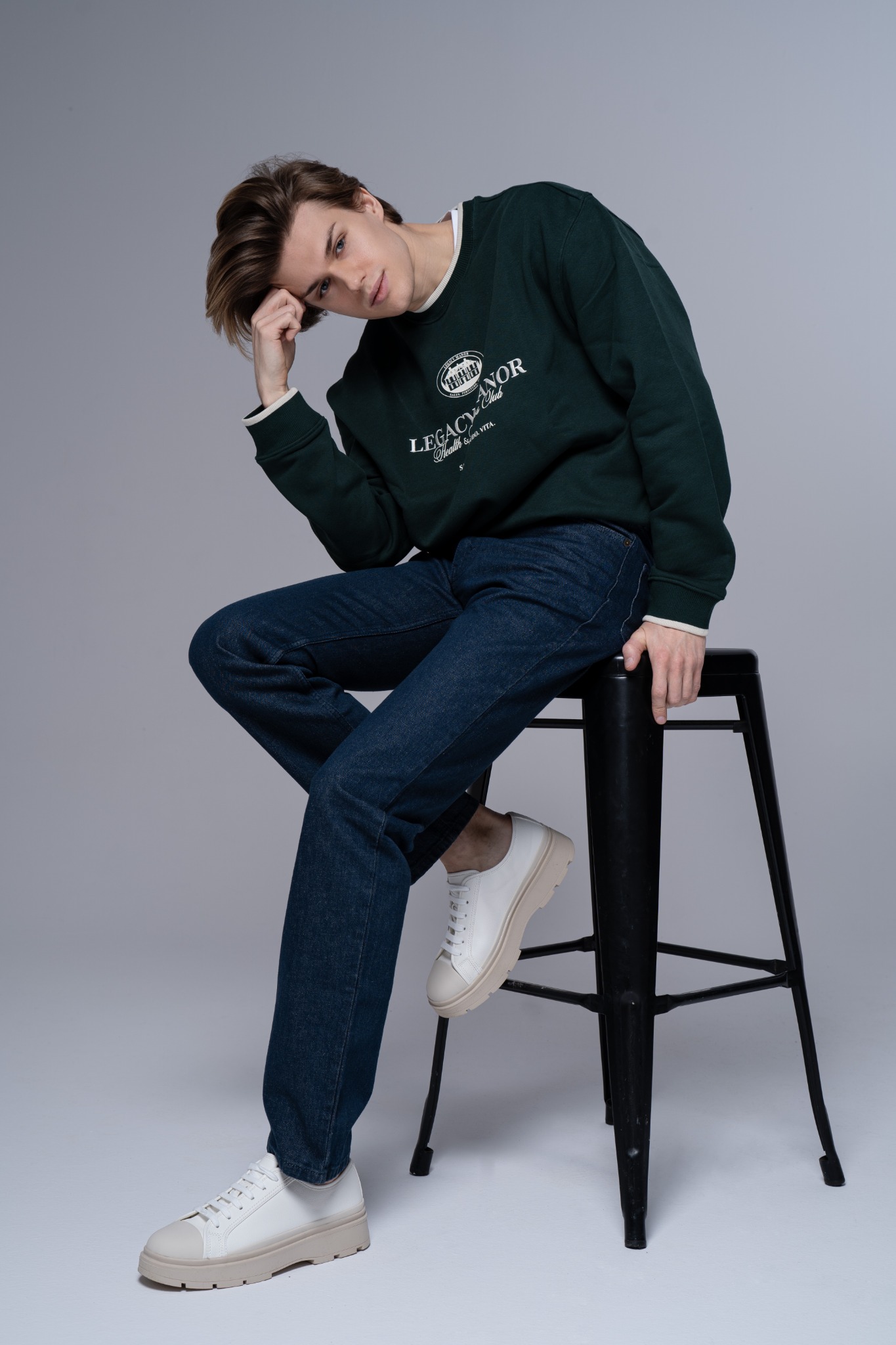 Młody mężczyzna w ciemnozielonej bluzie z logo, ciemnych jeansach i białych sneakersach, siedzący na czarnym stołku barowym na jednolitym szarym tle.