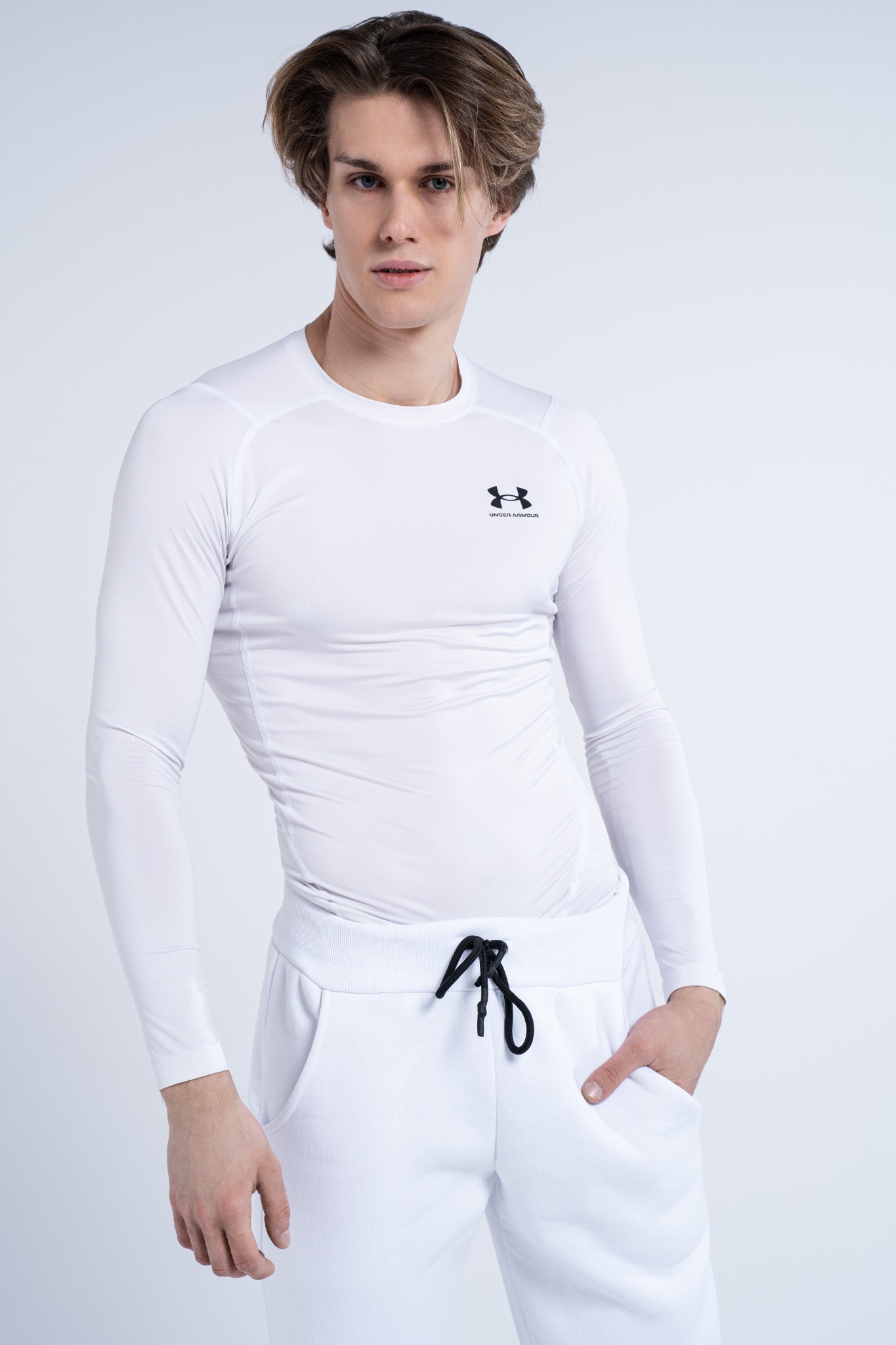 Młody mężczyzna w białym sportowym stroju Under Armour, pozujący na jasnym tle.