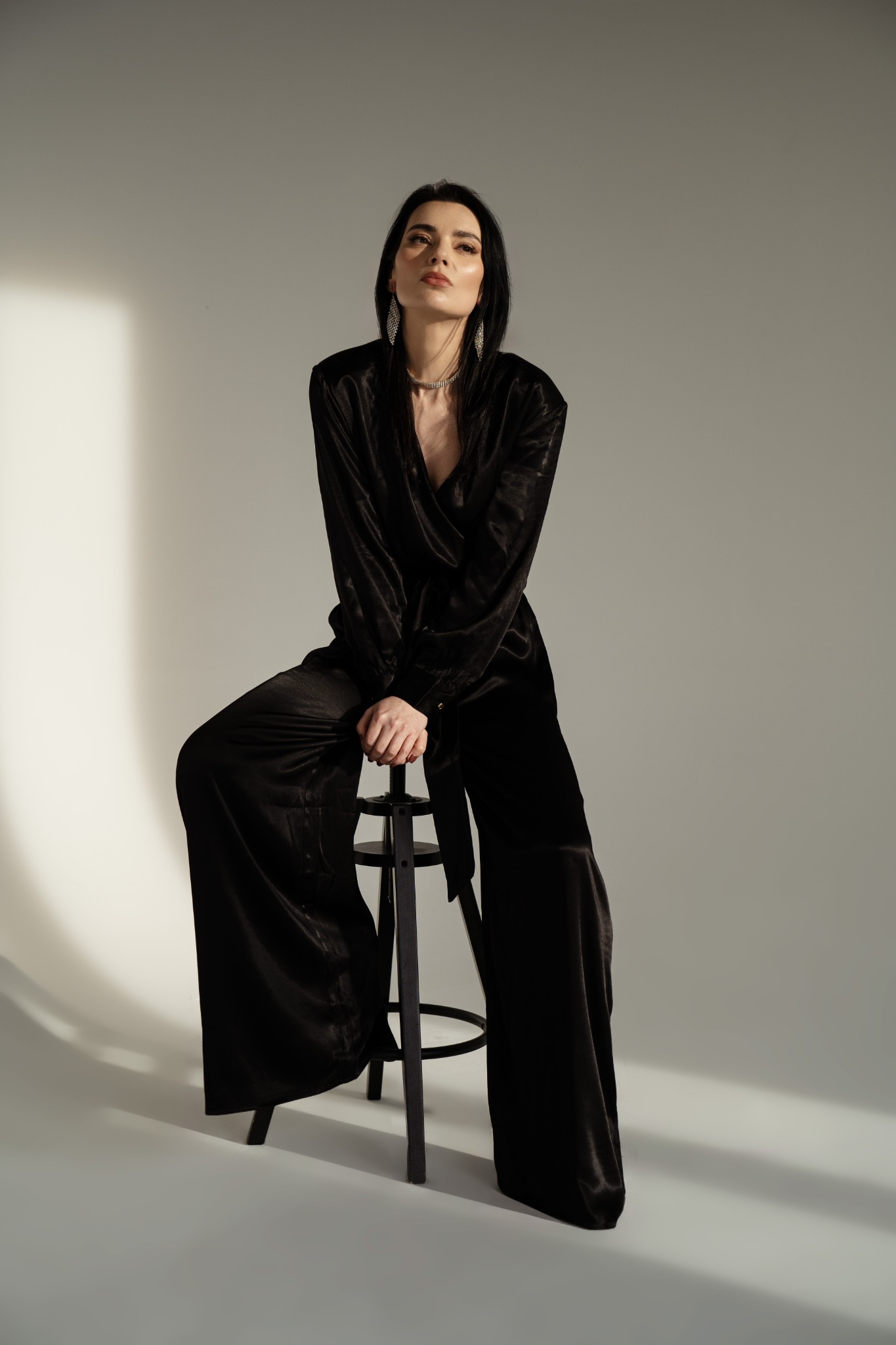 Portret kobiety w czarnym, satynowym kombinezonie siedzącej na czarnym stołku barowym, z delikatną biżuterią i spojrzeniem skierowanym w górę, na jasnym tle