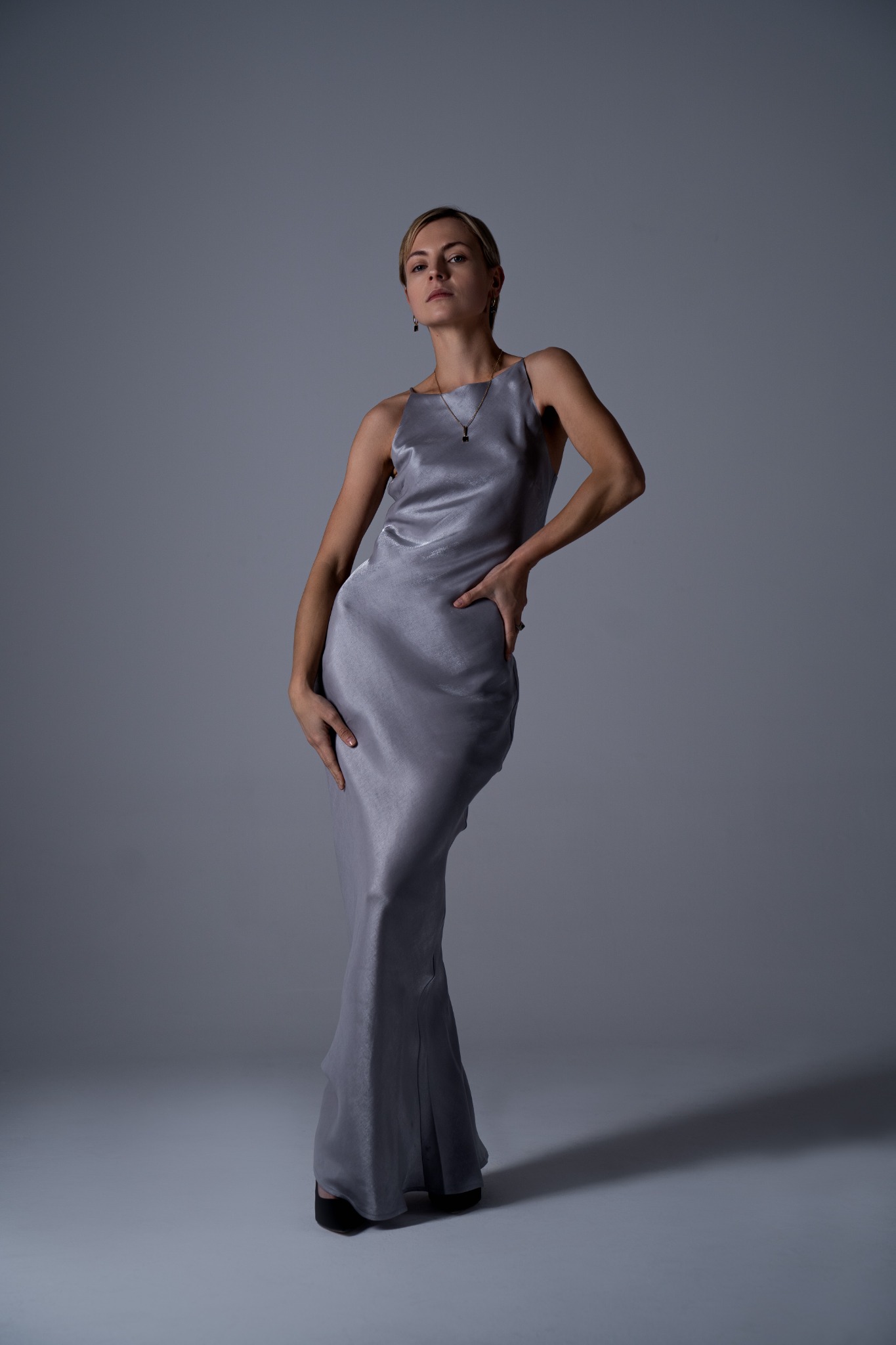 Portret kobiety w długiej, satynowej sukni w odcieniach szarości, pozującej na jednolitym, szarym tle. Kobieta ma krótkie blond włosy i delikatną biżuterię. Ujęcie studyjne, z subtelnym oświetleniem.