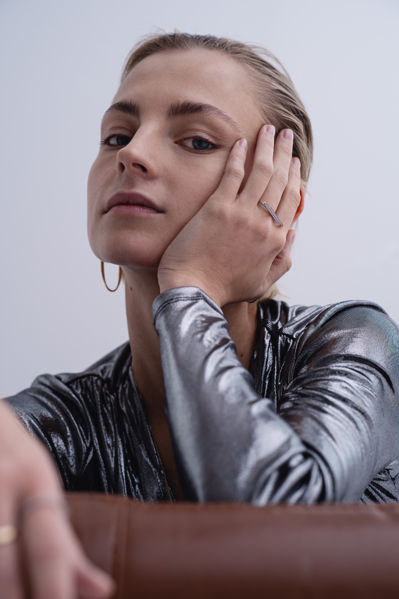 Portret kobiety w srebrnej bluzce, opierającej brodę na dłoni z pierścionkiem wysadzanym kamieniami, w minimalistycznym otoczeniu.