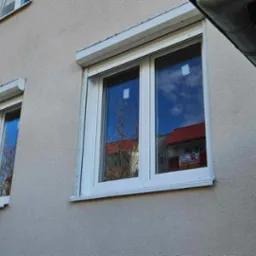 Białe okna z roletami zewnętrznymi zamontowane w budynku w Plewiskach, widoczne z zewnątrz, z odbiciem nieba i drzew w szybach.