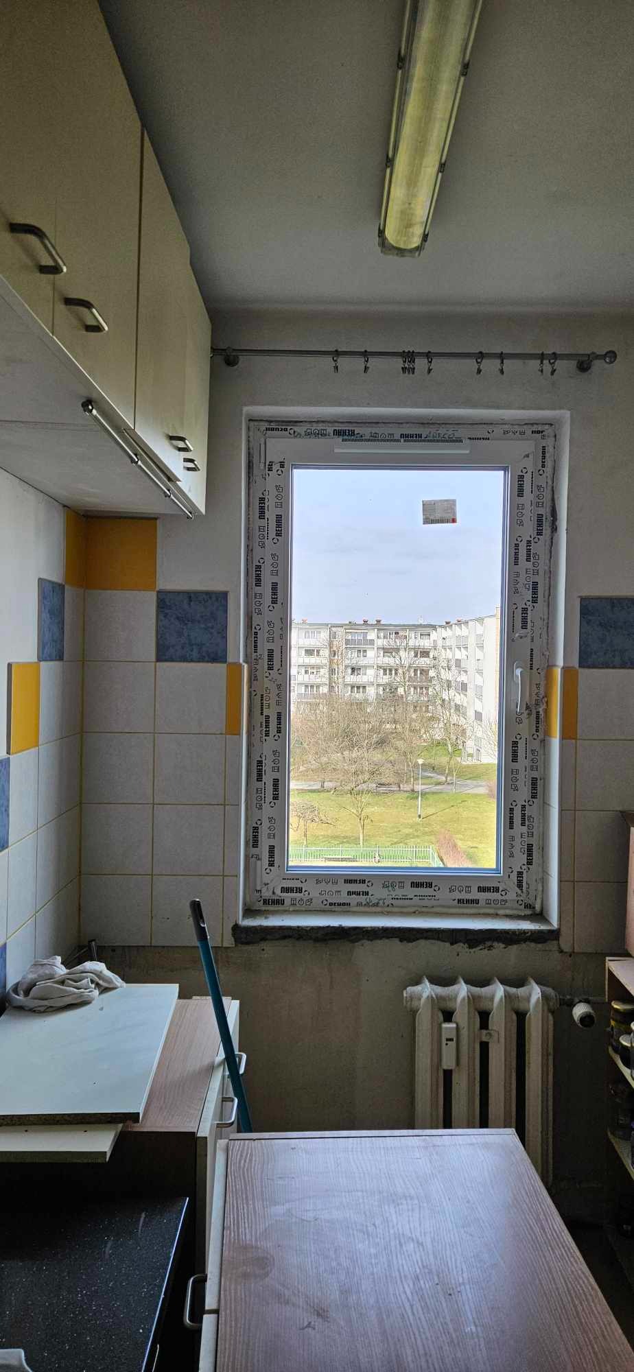 Wnętrze kuchni w trakcie remontu z widokiem na osiedle przez okno z ramą z tworzywa sztucznego, widoczne ślady po montażu, grzejnik żeliwny i fragmenty mebli.