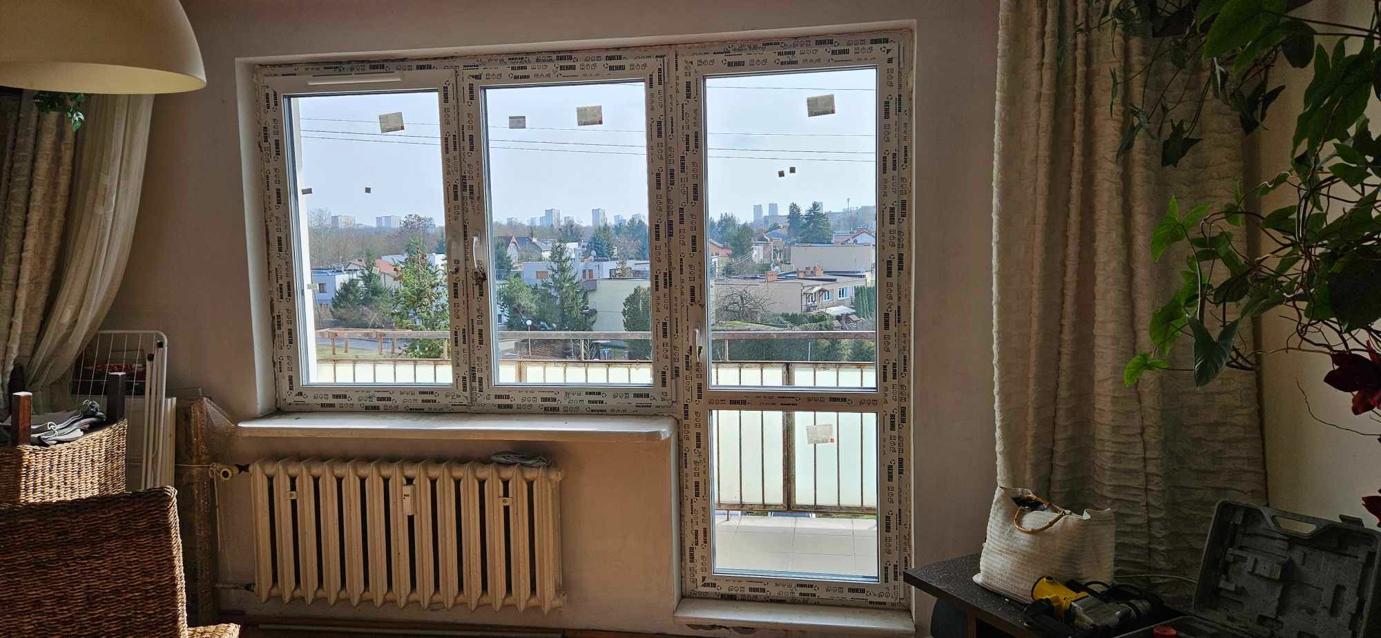 Nowo zamontowane, białe okna z PCV w mieszkaniu z widokiem na miasto, z jeszcze nieusuniętymi naklejkami producenta na szybach, widoczny grzejnik i narzędzia na stole.