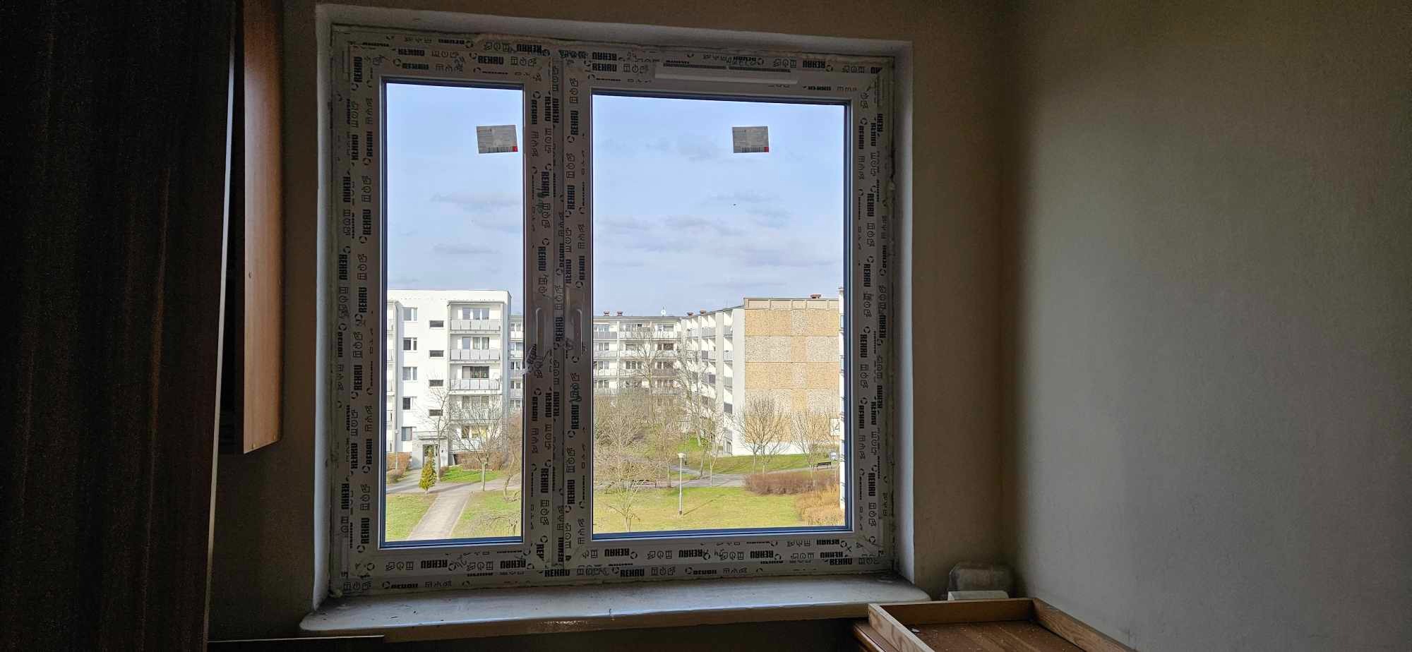 Nowe okno z białymi ramami z tworzywa sztucznego, widok na osiedle z zielenią, naklejki na szybach po montażu.