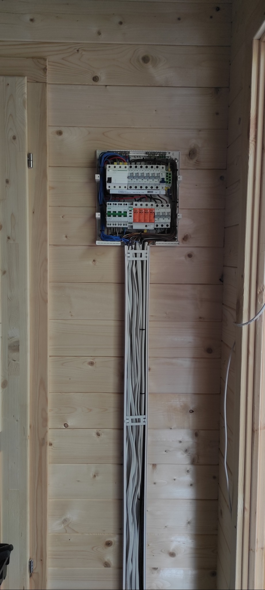 Rozdzielnia elektryczna z przewodami w korytku, zamontowana na ścianie z jasnego drewna wewnątrz budynku. Widoczne zabezpieczenia i okablowanie.