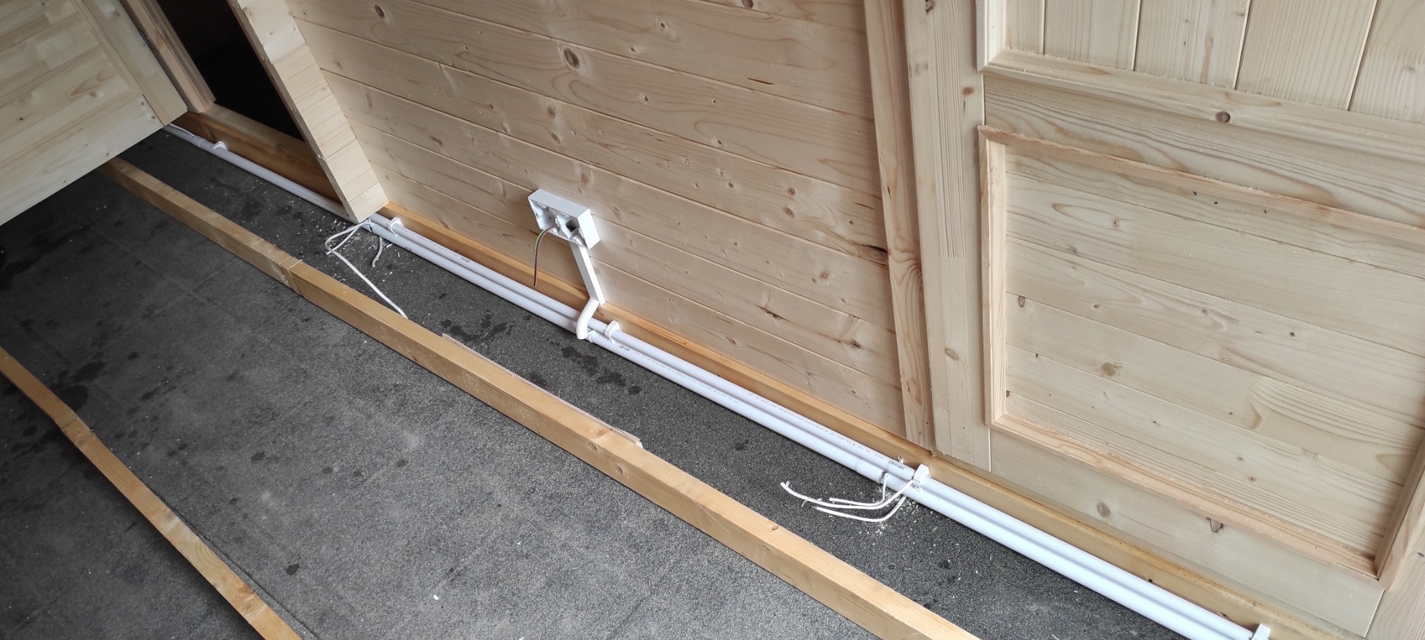 Wnętrze drewnianej konstrukcji w trakcie prac. Widoczne białe rury osłonowe na kable i puszka przyłączeniowa na ścianie z wystającymi przewodami.