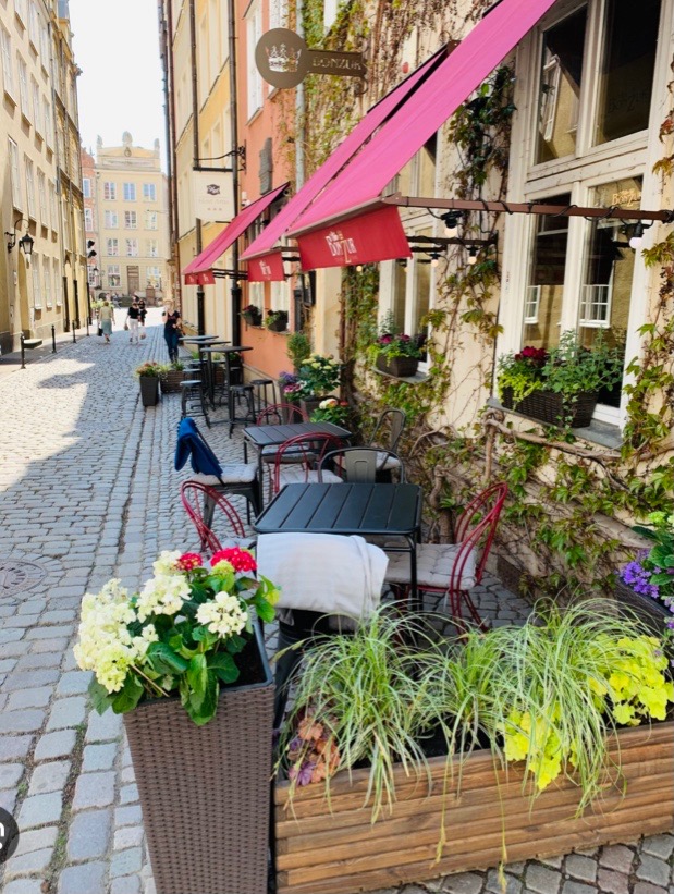Ogródek piwny restauracji Bonżur w Gdańsku z różowymi markizami, kwiatami w donicach i stolikami na brukowanej uliczce.