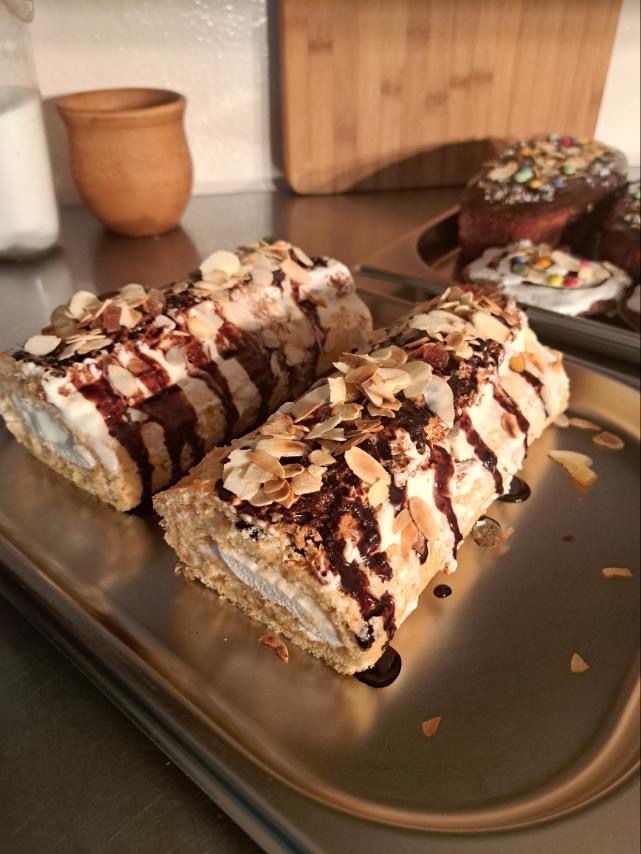 Dwa roladki biszkoptowe z kremem i polewą czekoladową, posypane płatkami migdałów, ułożone na srebrnej tacy, w tle babeczki wielkanocne z kolorową posypką.