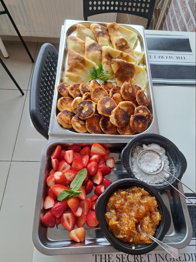 Metalowa taca z naleśnikami, serniczkami, pokrojonymi truskawkami, konfiturą i cukrem pudrem, udekorowana listkami mięty, na stole z gazetą w tle.