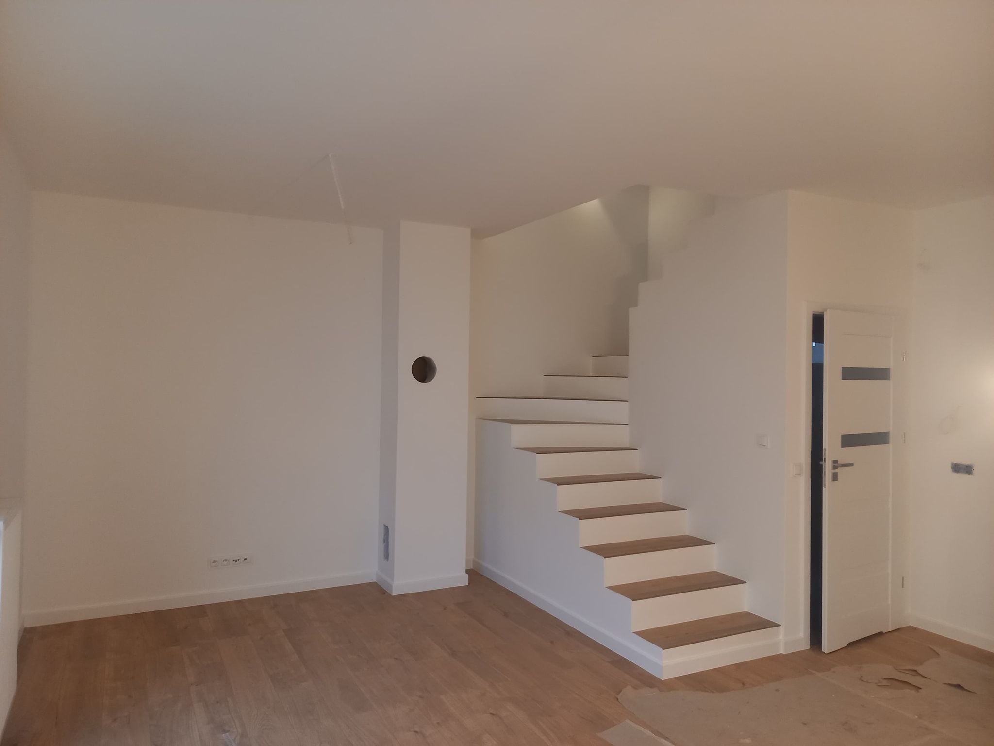 Jasne wnętrze z drewnianą podłogą i białymi schodami bez poręczy, częściowo odsłonięte drzwi wewnętrzne, widoczne gniazdka elektryczne i otwór w ścianie.