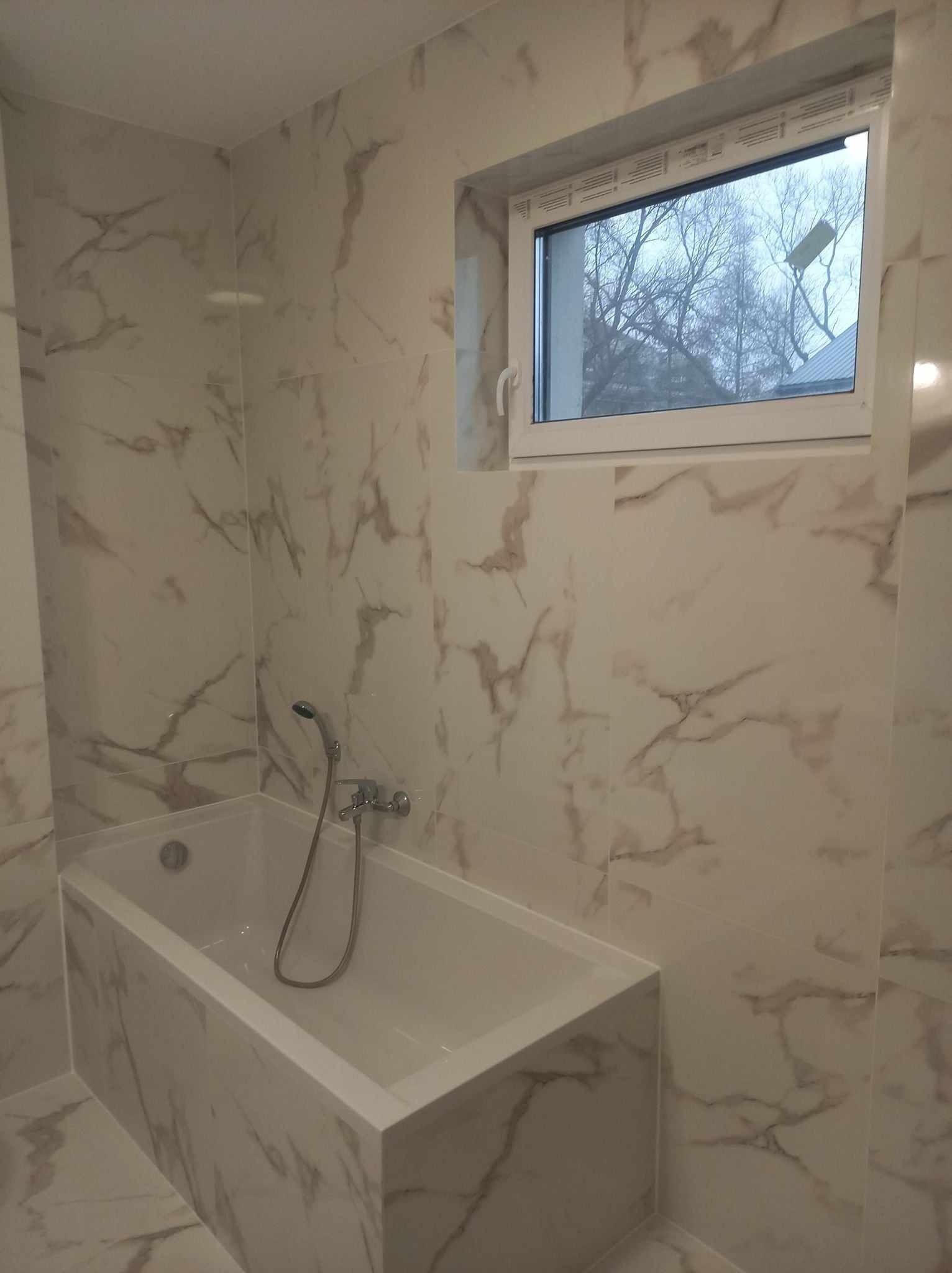 Nowoczesna łazienka z wanną obudowaną płytkami imitującymi marmur, białe okno z widokiem na drzewa bez liści.