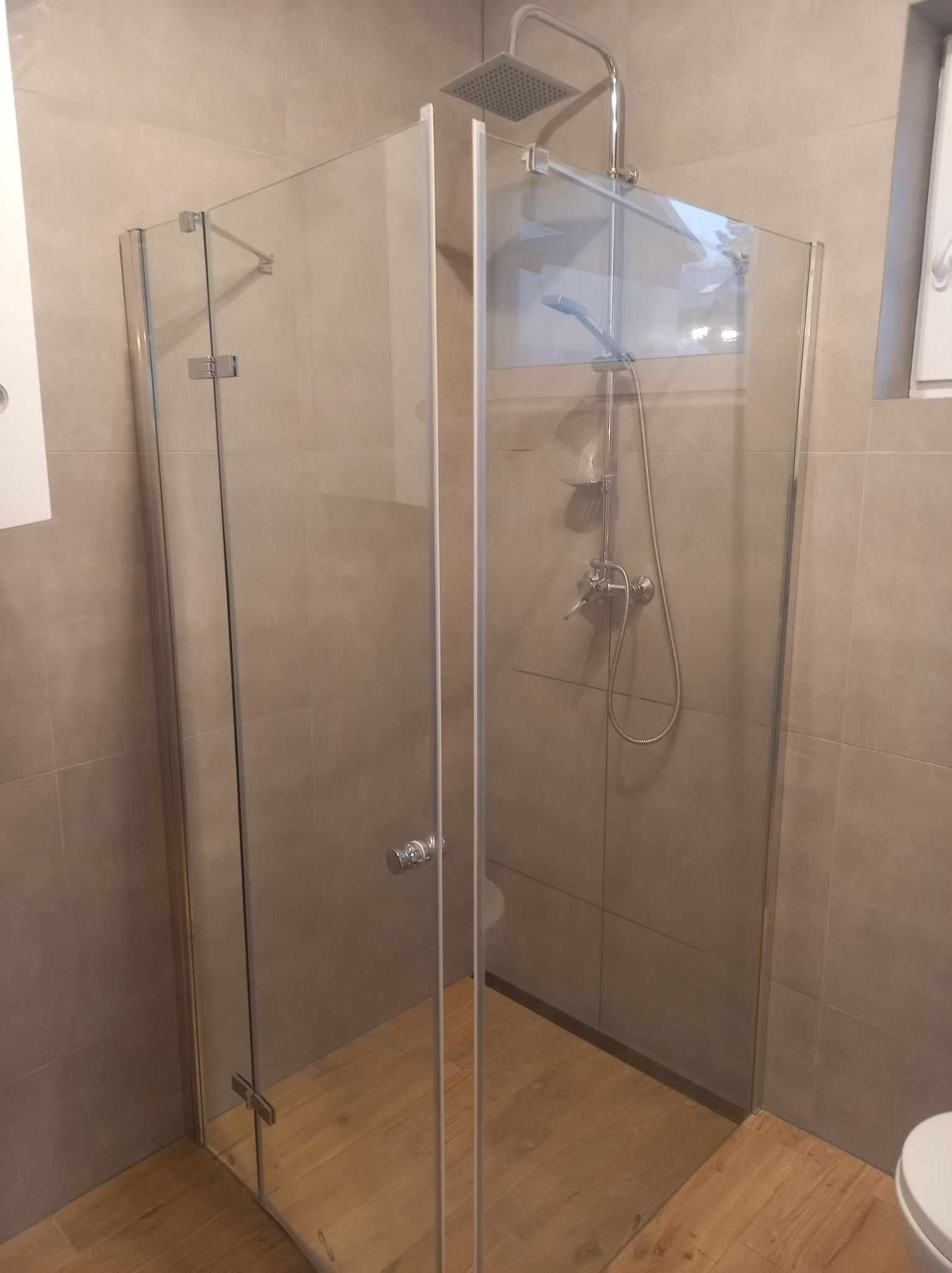 Nowoczesna kabina prysznicowa ze szklanymi ściankami, chromowaną armaturą i deszczownicą, drewnopodobna podłoga, szare płytki ścienne.