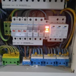 Rozdzielnia elektryczna Hager z widocznymi bezpiecznikami, SPD i listwami zaciskowymi, z podłączonymi przewodami o różnych kolorach i oznaczeniach, z zapaloną diodą na jednym z ograniczników przepięć.