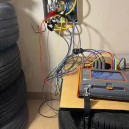 Elektryk w Katowicach wykonuje pomiary w skrzynce elektrycznej za pomocą miernika Sonel, widoczne przewody i stos opon w tle.