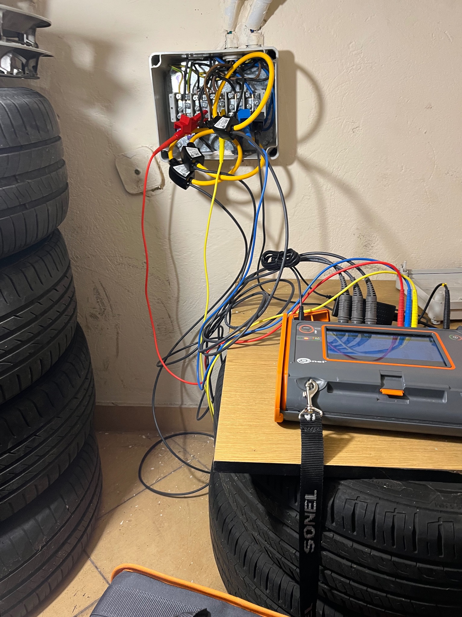 Elektryk w Katowicach wykonuje pomiary w skrzynce elektrycznej za pomocą miernika Sonel, widoczne przewody i stos opon w tle.