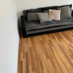 Szary narożnik z poduszkami w odcieniach szarości i pudrowego r&oacute;żu na tle drewnianej podłogi i białej ściany.