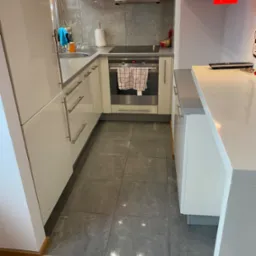 Wnętrze nowoczesnej kuchni z białymi szafkami, czerwonymi górnymi szafkami, szarymi płytkami podłogowymi i drewnianą podłogą w przejściu, z widocznym piekarnikiem i akcesoriami kuchennymi.