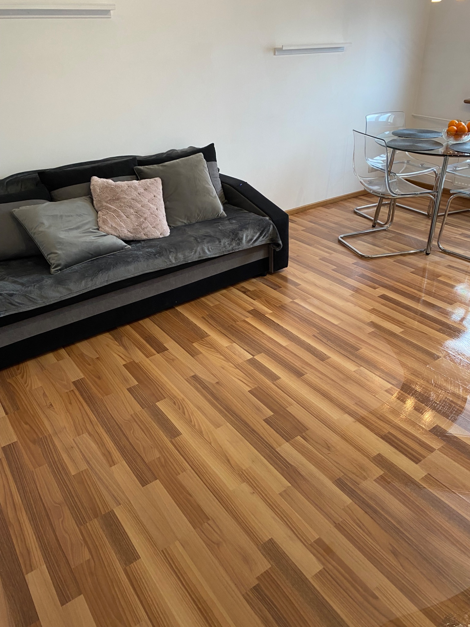 Błyszcząca, świeżo umyta podłoga panelowa z widocznym odbiciem światła, obok czarna sofa z szarymi i różową poduszką oraz szklany stół z krzesłami w tle.