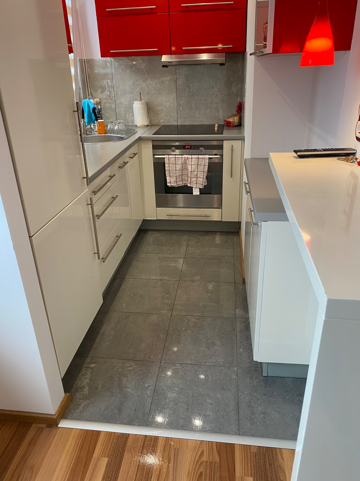 Wnętrze nowoczesnej kuchni z białymi szafkami, czerwonymi górnymi szafkami, szarymi płytkami podłogowymi i drewnianą podłogą w przejściu, z widocznym piekarnikiem i akcesoriami kuchennymi.