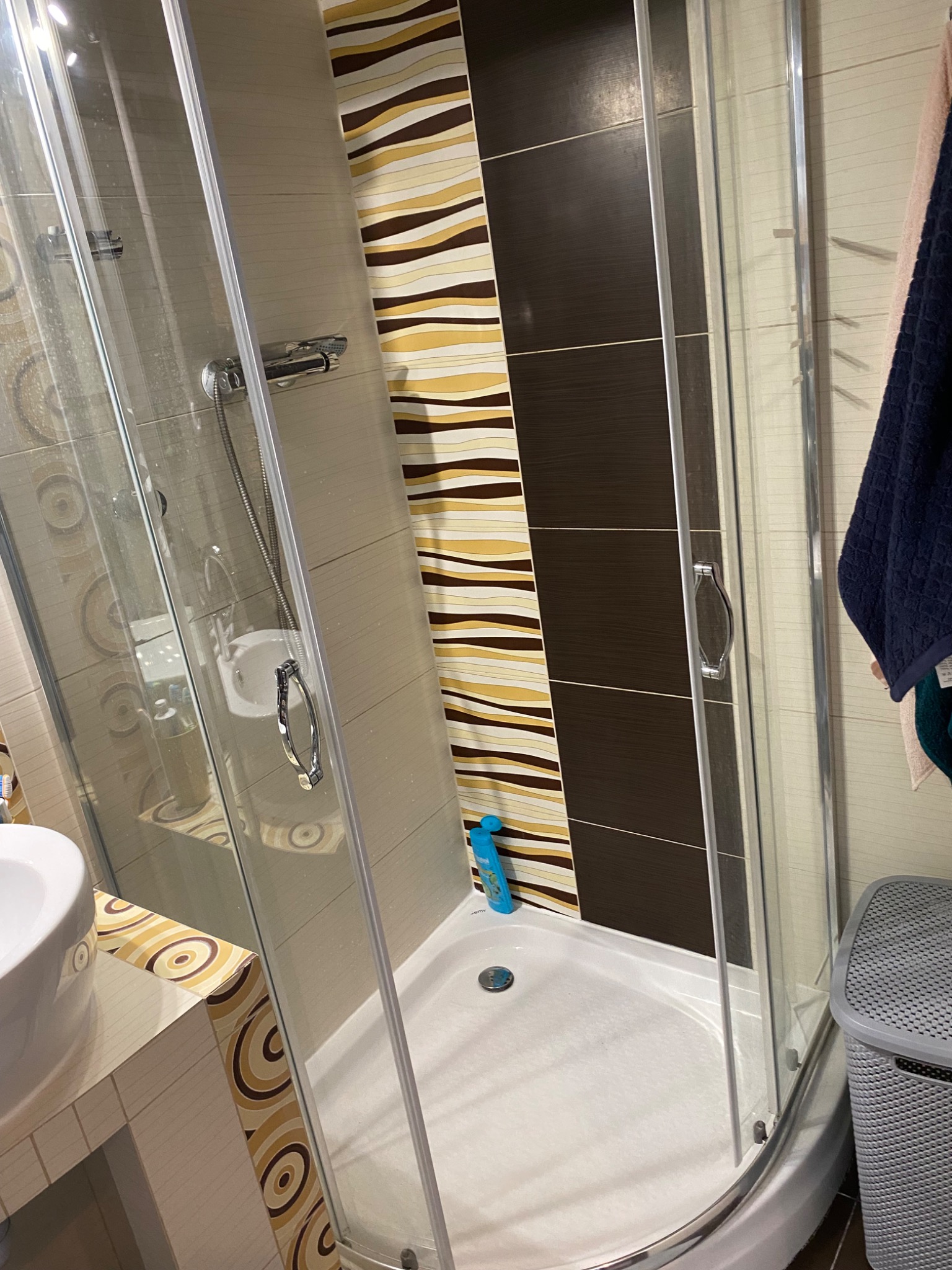 Nowoczesna kabina prysznicowa z przesuwnymi drzwiami, widoczna butelka płynu do mycia pod prysznicem, dekoracyjne płytki w odcieniach brązu i beżu.