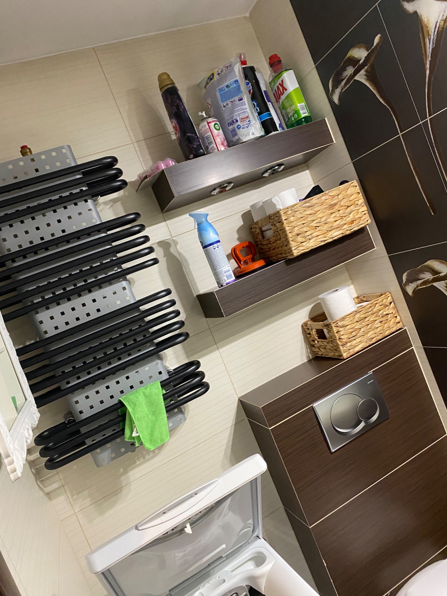 Toaleta z podwieszanym stelażem, koszyki wiklinowe na półkach z zapasem papieru toaletowego, środki czystości i grzejnik drabinkowy z zieloną ściereczką w nowoczesnej łazience.