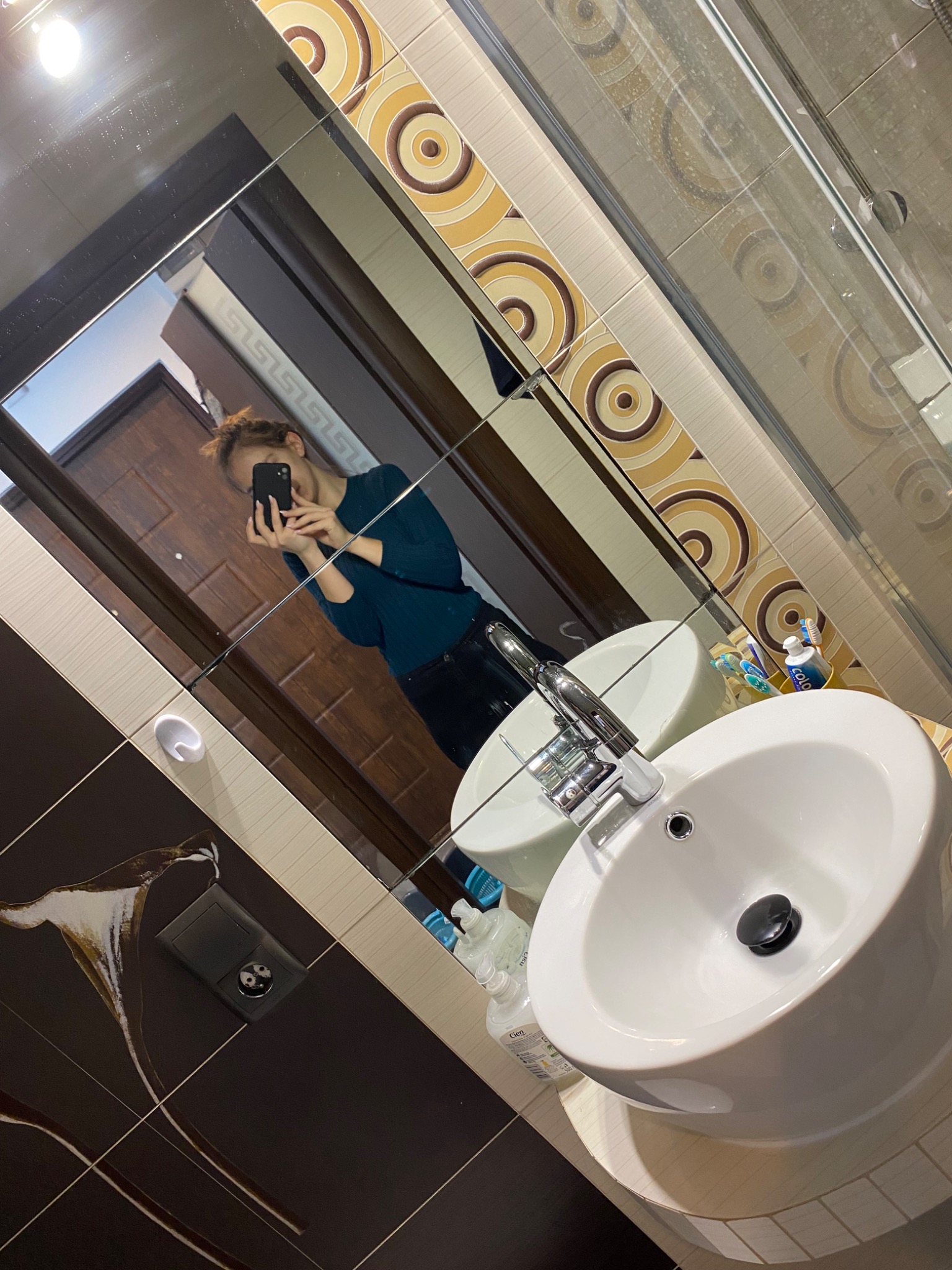 Umywalka w łazience z baterią i odbiciem osoby robiącej zdjęcie w lustrze. Widoczne płytki z wzorem i akcesoria łazienkowe.