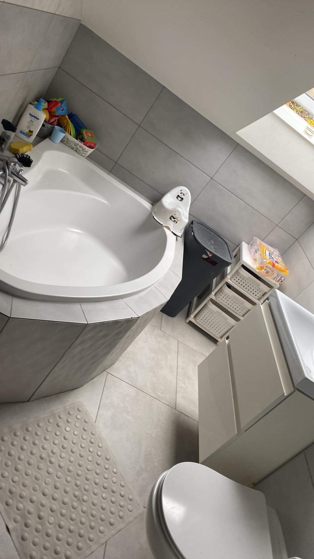 Widok z góry na czystą, białą wannę narożną z szarymi płytkami, białą matą antypoślizgową i akcesoriami łazienkowymi, w jasnej łazience z oknem dachowym.