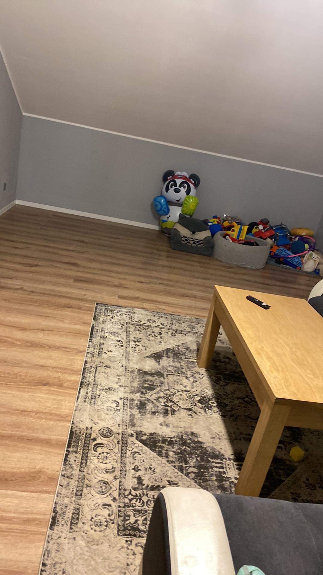 Pokój dziecięcy z drewnianą podłogą, dywanem w stylu vintage, drewnianym stolikiem, fotelikiem i stertą zabawek oraz balonem w kształcie pandy przy ścianie.