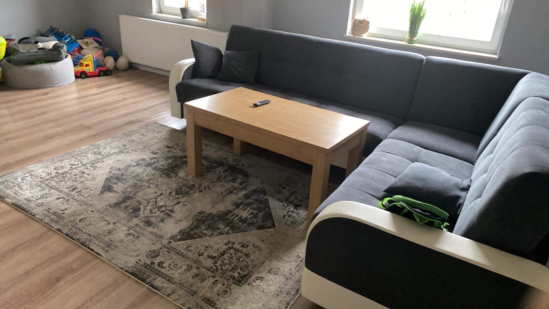 Salon z szarą narożną sofą, drewnianym stolikiem kawowym z pilotem, wzorzystym dywanem i zabawkami w rogu pokoju, oświetlony przez okno.