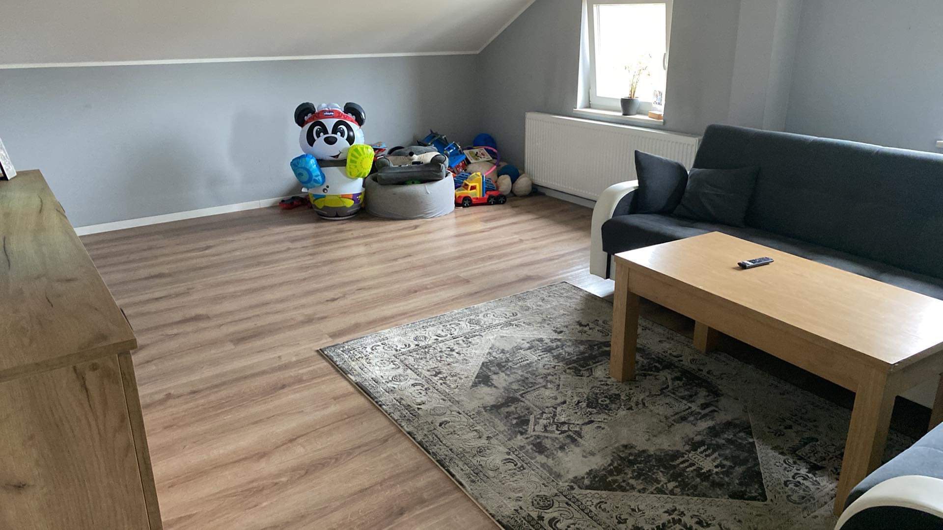 Pokój na poddaszu z drewnianą podłogą, szarą ścianą, sofą, stolikiem kawowym z pilotem, dywanem w stylu vintage i zabawkami w rogu; widok ogólny.