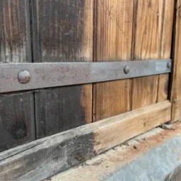 Fragment drewnianej, zabytkowej bramy z metalowym okuciem, widoczne ślady renowacji drewna i rdzy na metalu.