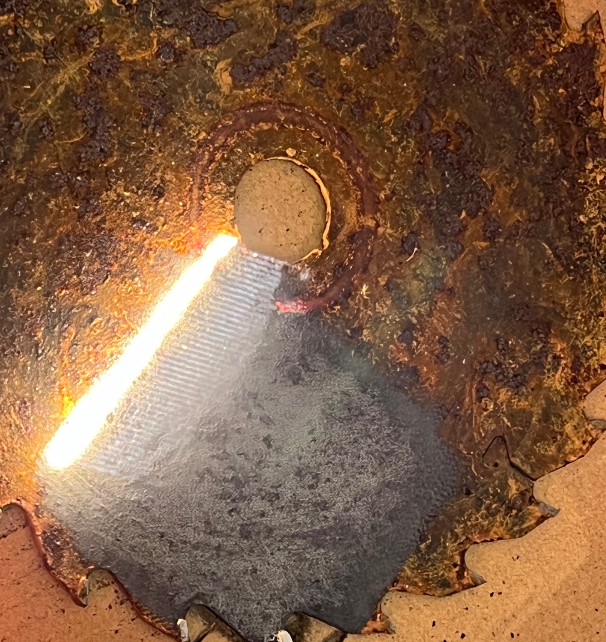 Silnie skorodowana, okrągła metalowa tarcza z centralnym otworem i fragmentami ostrzy, o nierównomiernej powierzchni z widocznymi śladami rdzy i przebarwień, oświetlona z boku jasnym światłem.