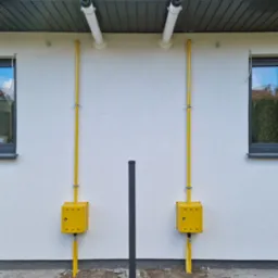 Żółte rury gazowe biegnące wzdłuż białej ściany budynku, z odpowietrznikami pod dachem i skrzynkami przy ziemi, widoczne okna z szarymi ramami i czarny słupek w pobliżu.
