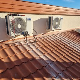 Firma Usługowa Natalia Wróblewska - Dwa zamontowane na elewacji budynku klimatyzatory AUX DC Inverter, widoczne narzędzia pomiarowe podłączone do jednego z urządzeń, dachówka ceramiczna w tle.