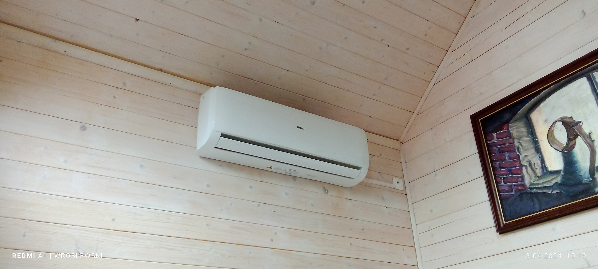 Biały klimatyzator Haier zamontowany na ścianie z jasnych, drewnianych paneli boazeryjnych w domku, obok wisi obraz w ciemnej ramie.