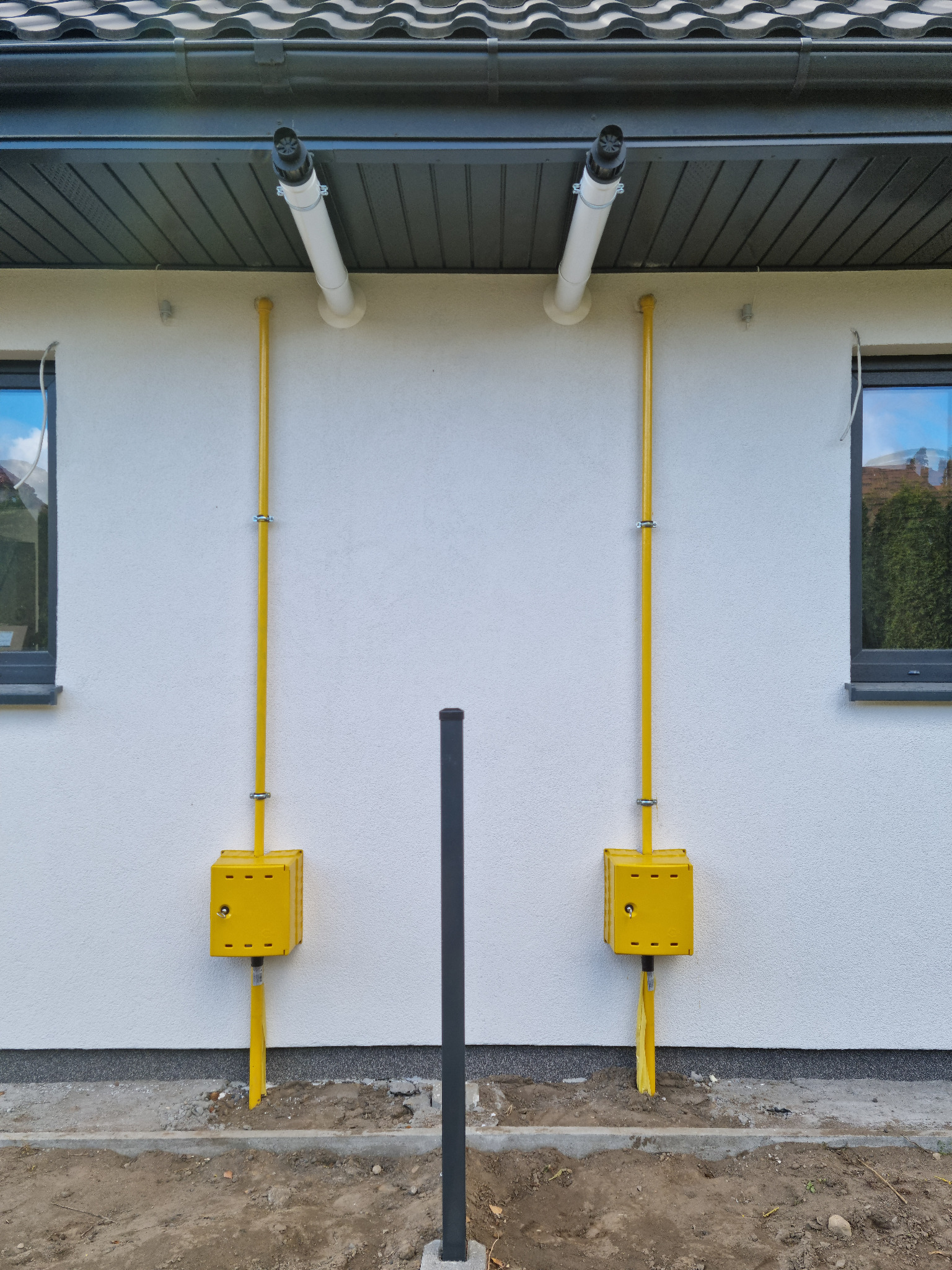 Żółte rury gazowe biegnące wzdłuż białej ściany budynku, z odpowietrznikami pod dachem i skrzynkami przy ziemi, widoczne okna z szarymi ramami i czarny słupek w pobliżu.
