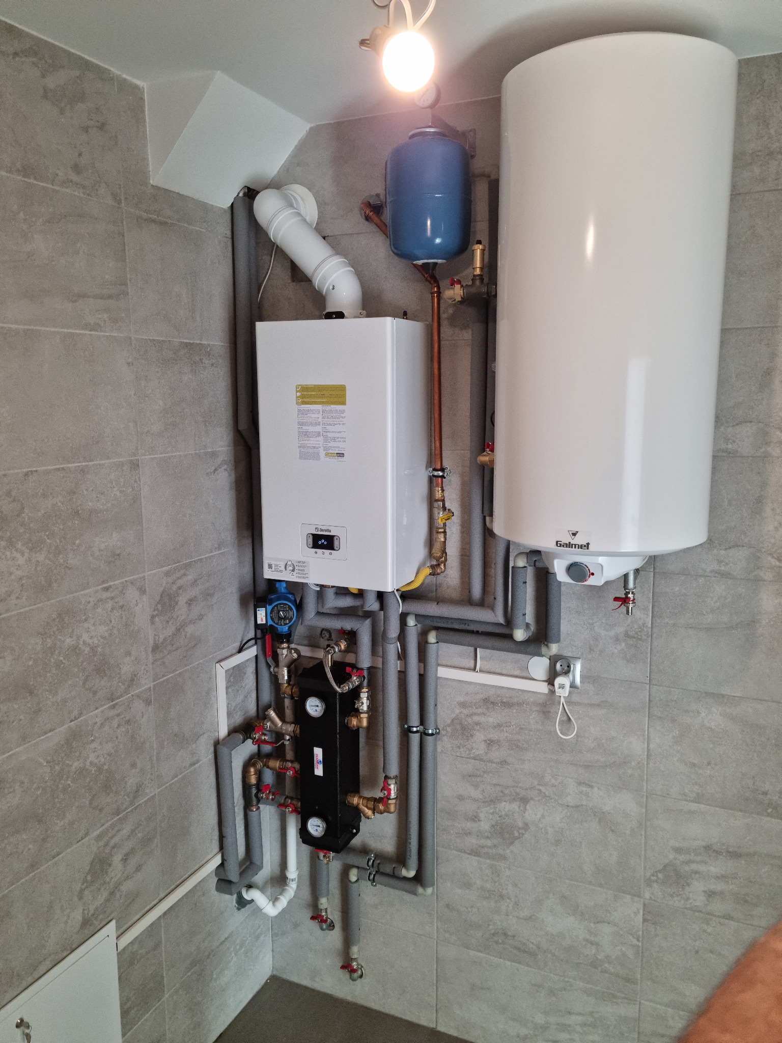 Nowoczesna kotłownia z piecem gazowym, zasobnikiem ciepłej wody i pompą obiegową, całość na tle szarych płytek ceramicznych.