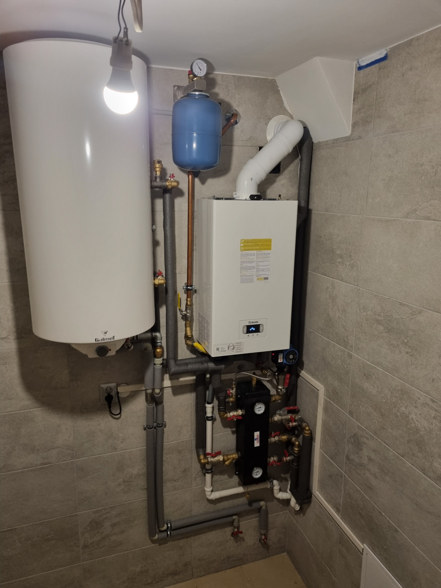 Zestaw instalacji grzewczej z piecem gazowym Beretta i zasobnikiem Galmet, rury w szarej izolacji, manometr na niebieskim naczyniu wzbiorczym, płytki imitujące kamień na ścianach.