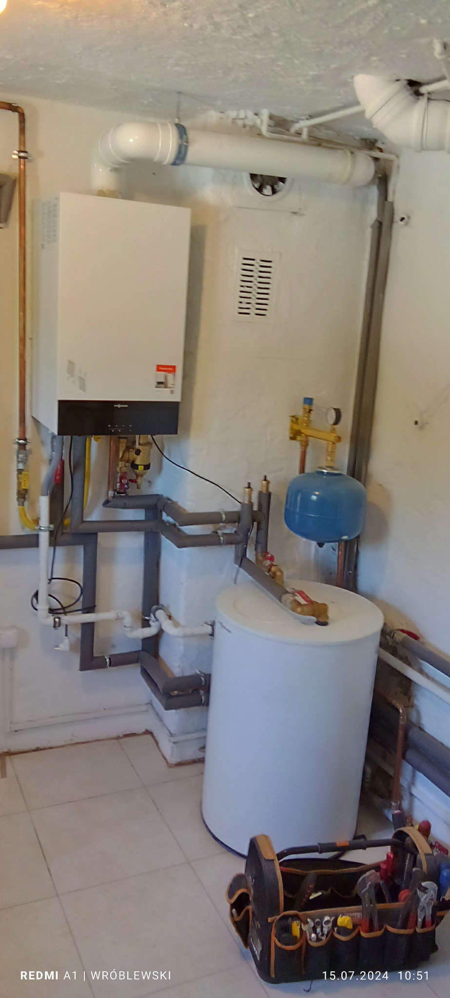 Biały piec gazowy Viessmann zamontowany na ścianie w piwnicy, z widocznymi rurami miedzianymi i szarymi, zbiornik wody oraz skrzynka z narzędziami hydraulika na podłodze.