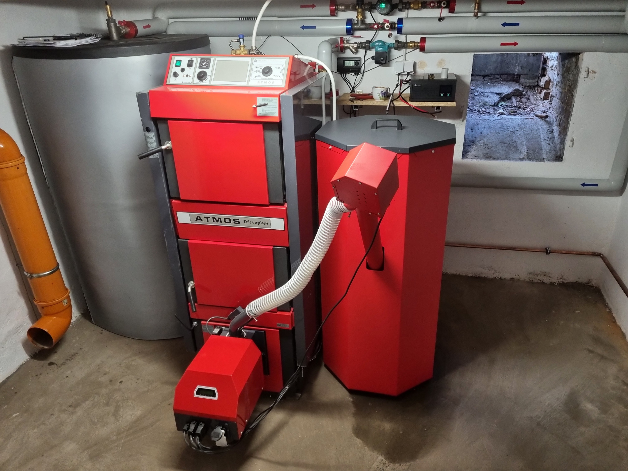 Czerwony piec na paliwo stałe Atmos Drevoplyn z zasobnikiem w kotłowni, widoczne rury instalacji grzewczej i otwór wentylacyjny w ścianie.