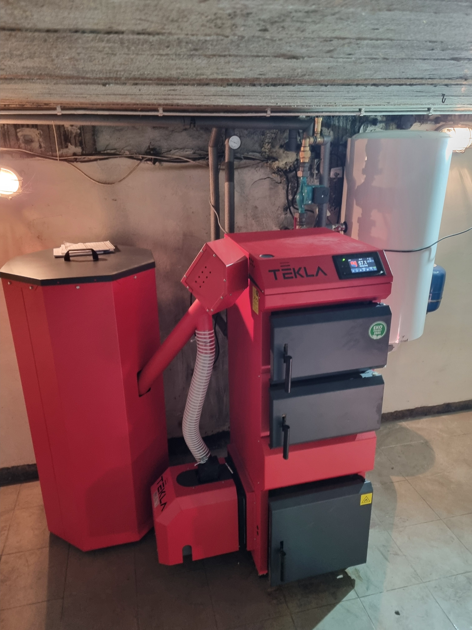 Czerwony piec Tekla z podajnikiem, widoczny w piwnicy z instalacją hydrauliczną i zasobnikiem ciepłej wody.