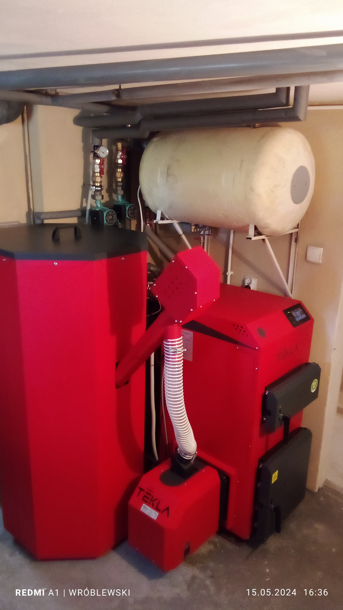 Czerwony piec na paliwo stałe Tekla z zasobnikiem, biały zbiornik akumulacyjny u góry, rury i zawory w kotłowni.
