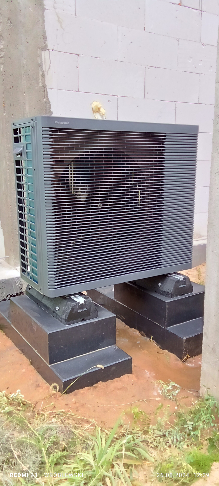 Zewnętrzna jednostka klimatyzacji Panasonic zamontowana na betonowych podstawach, widoczne ślady wilgoci na ziemi.