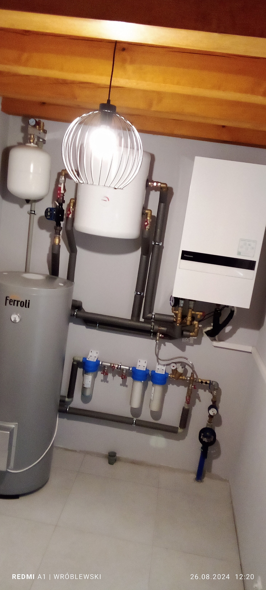 Zestaw instalacji grzewczej z szarym zasobnikiem Ferroli, białym zbiornikiem wyrównawczym, piecem gazowym i filtrami wody, oświetlony dekoracyjną lampą z żarówką.